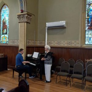 A man playing the piano in church while a woman stands beside the piano.